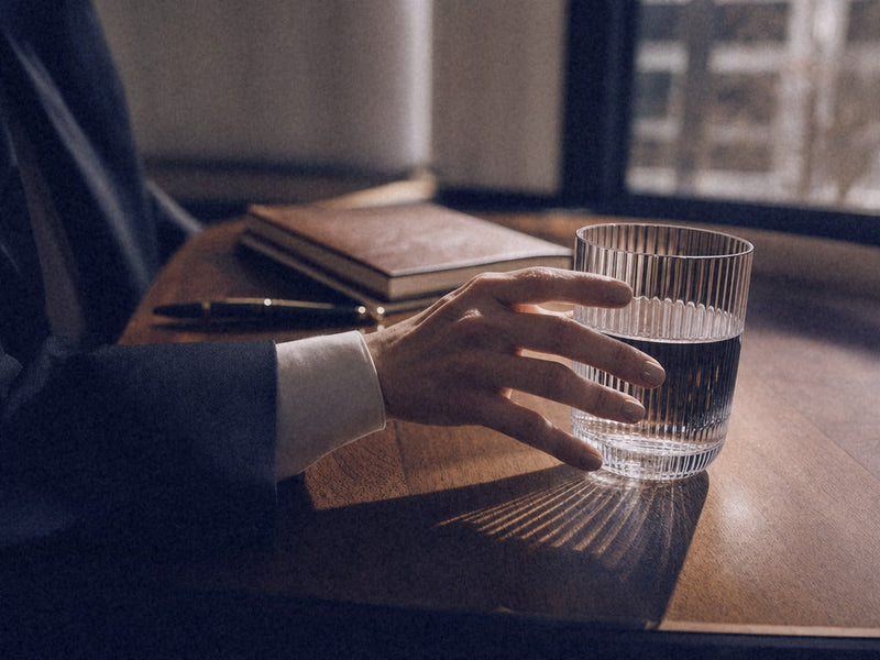 Close-up of a hand in a white shirt cuff and dark blazer reaching for a ribbed glass of water on a walnut desk, a large window and a tree behind, afternoon light.