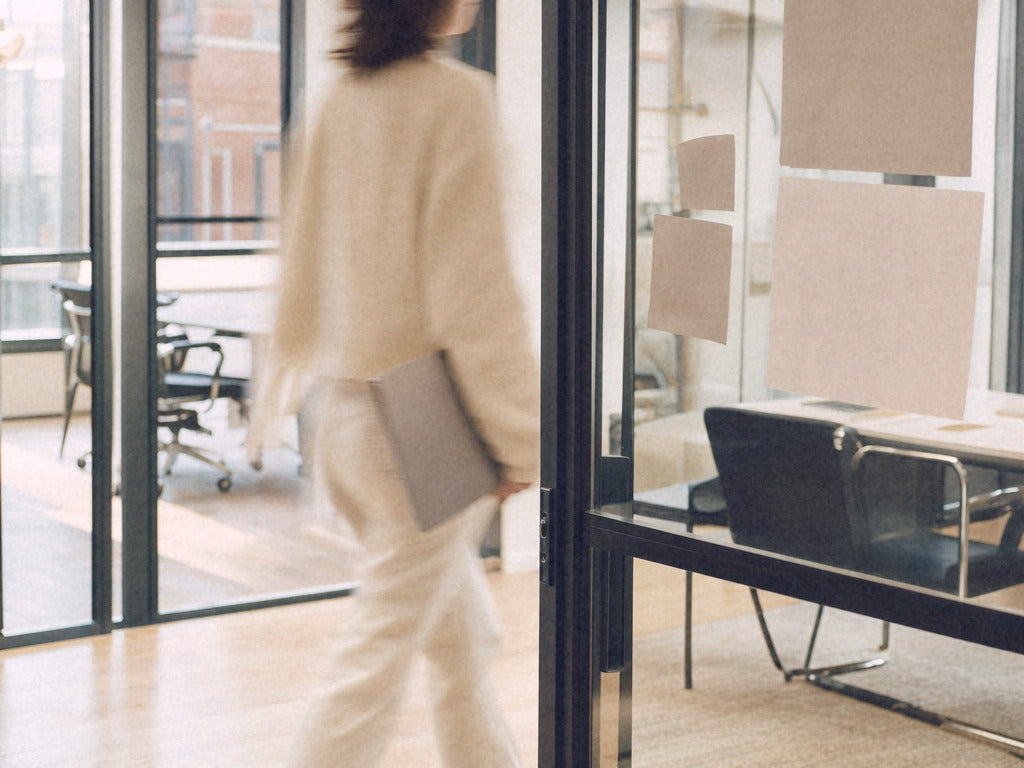 A person in a cream sweater hurrying between two glass-walled meeting rooms in a bright Montreal office, softly motion-blurred.