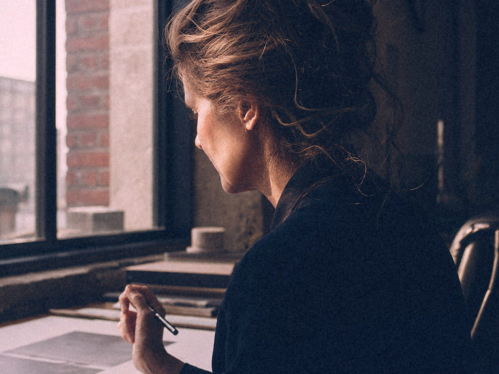 A woman seen from behind at a desk by a tall window with red brick outside, pen in hand, absorbed in a page in morning light.