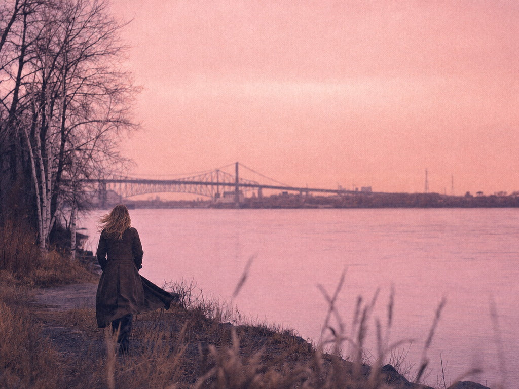 A woman walks along the river at dawn, hair lifting in the wind.